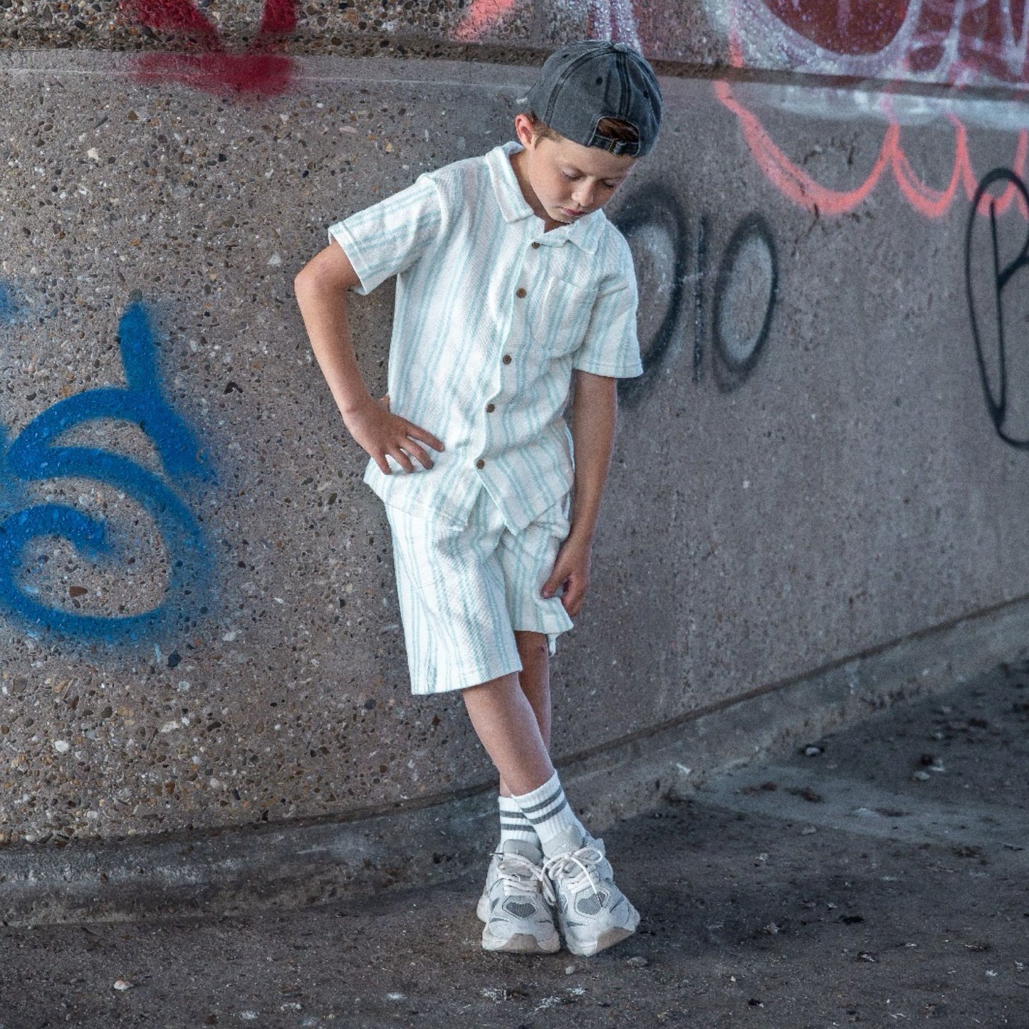 Kind met een grijs gewassen denim pet op achterstevoren, van voren  gefotografeerd in een urban omgeving met graffiti.